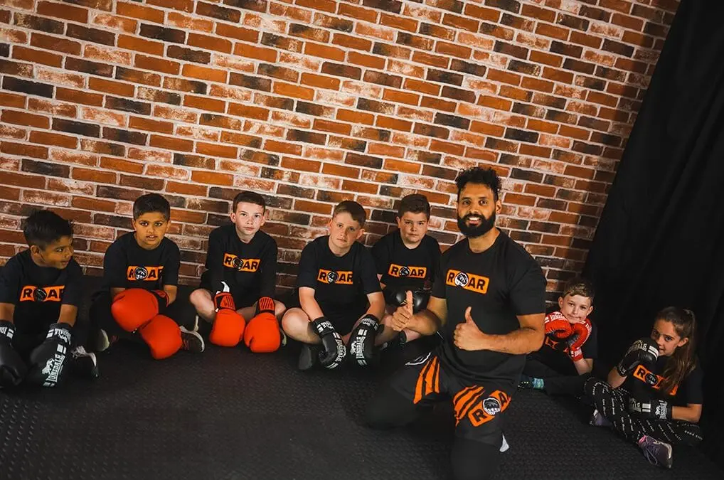 Coach poses with group of young boxers wearing gloves, seated together at Roar Martial Arts