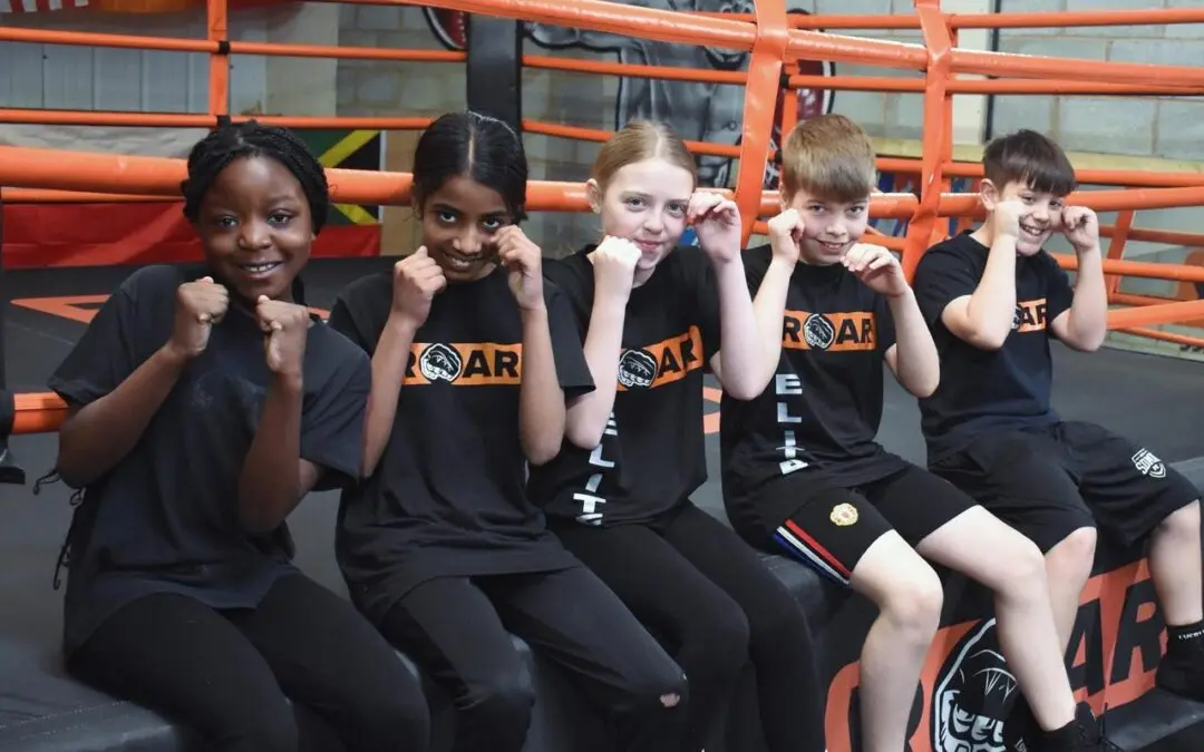 Group of smiling kids wearing Roar Martial Arts shirts posing in boxing stances by the ring